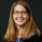 This is a headshot of a person with long brown hair and glasses, smiling, against a dark background.